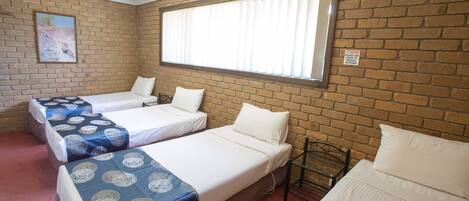 A room with three beds arranged in a row against a brick wall, a window with blinds, and a painting on the wall.