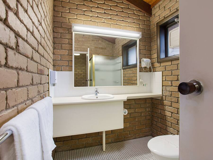 A bathroom with a white sink, a mirror, and a toilet. The walls are made of brick, and there is a window above the toilet.
