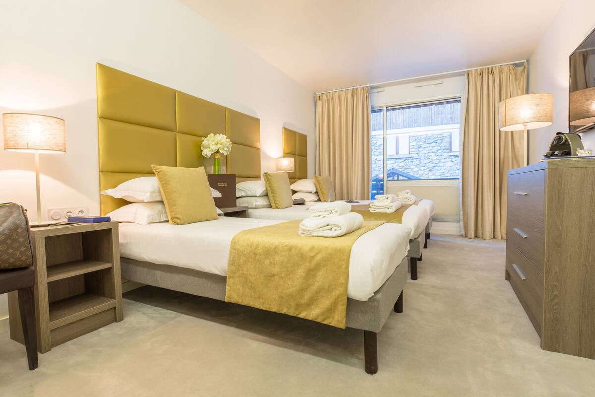 Habitación de hotel con dos camas, una mesita de noche de madera, un aparador y un amplio ventanal con cortinas.