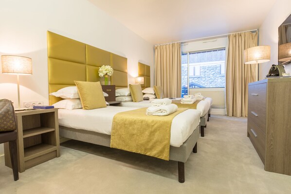 A hotel room with two beds, a wooden nightstand, a dresser, and a large window with curtains. - Madame Vacances Hôtel Courchevel Olympic (Courchevel)