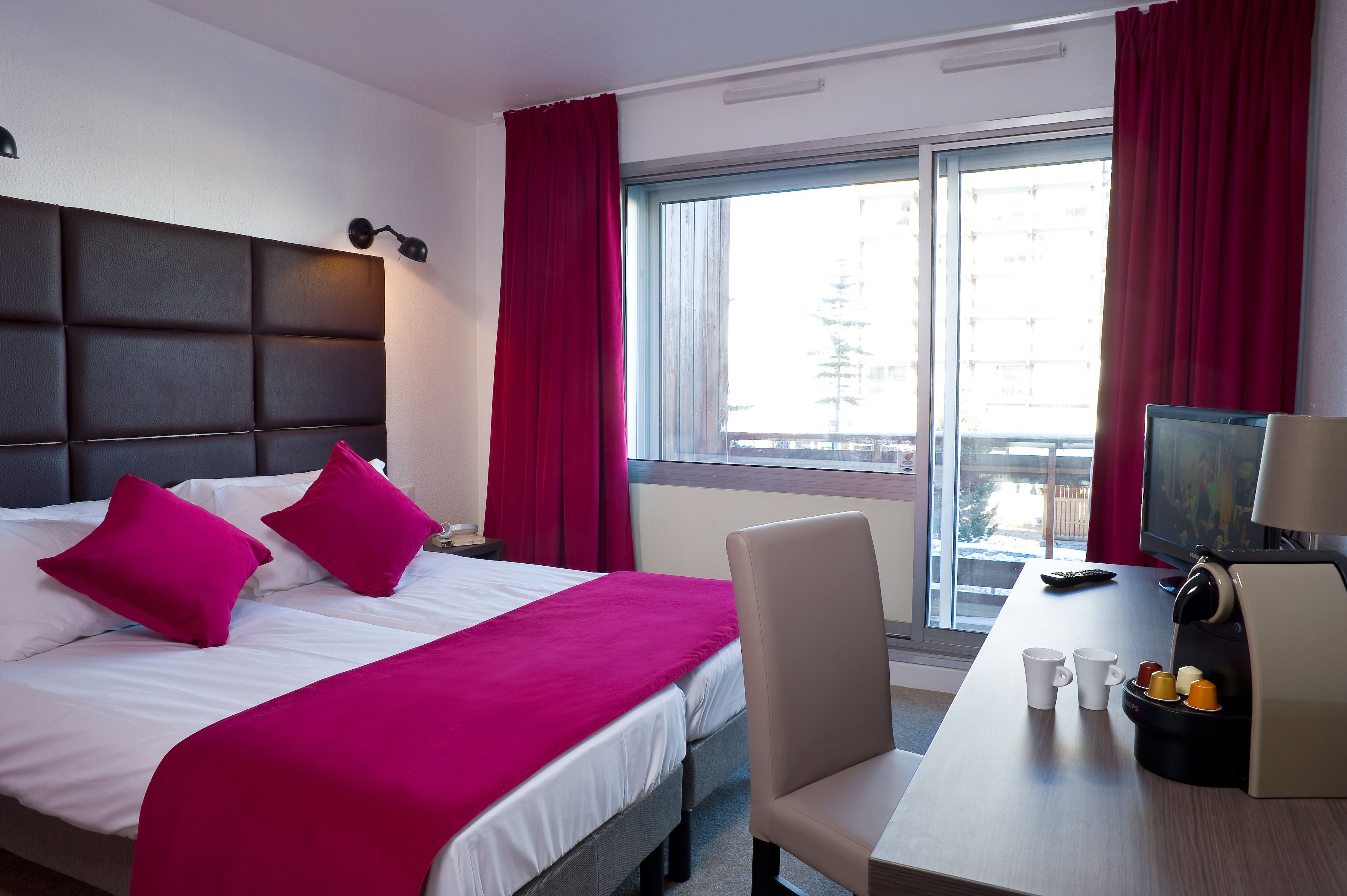 A hotel room with a bed, a desk with a coffee maker, a TV, and a window with curtains.