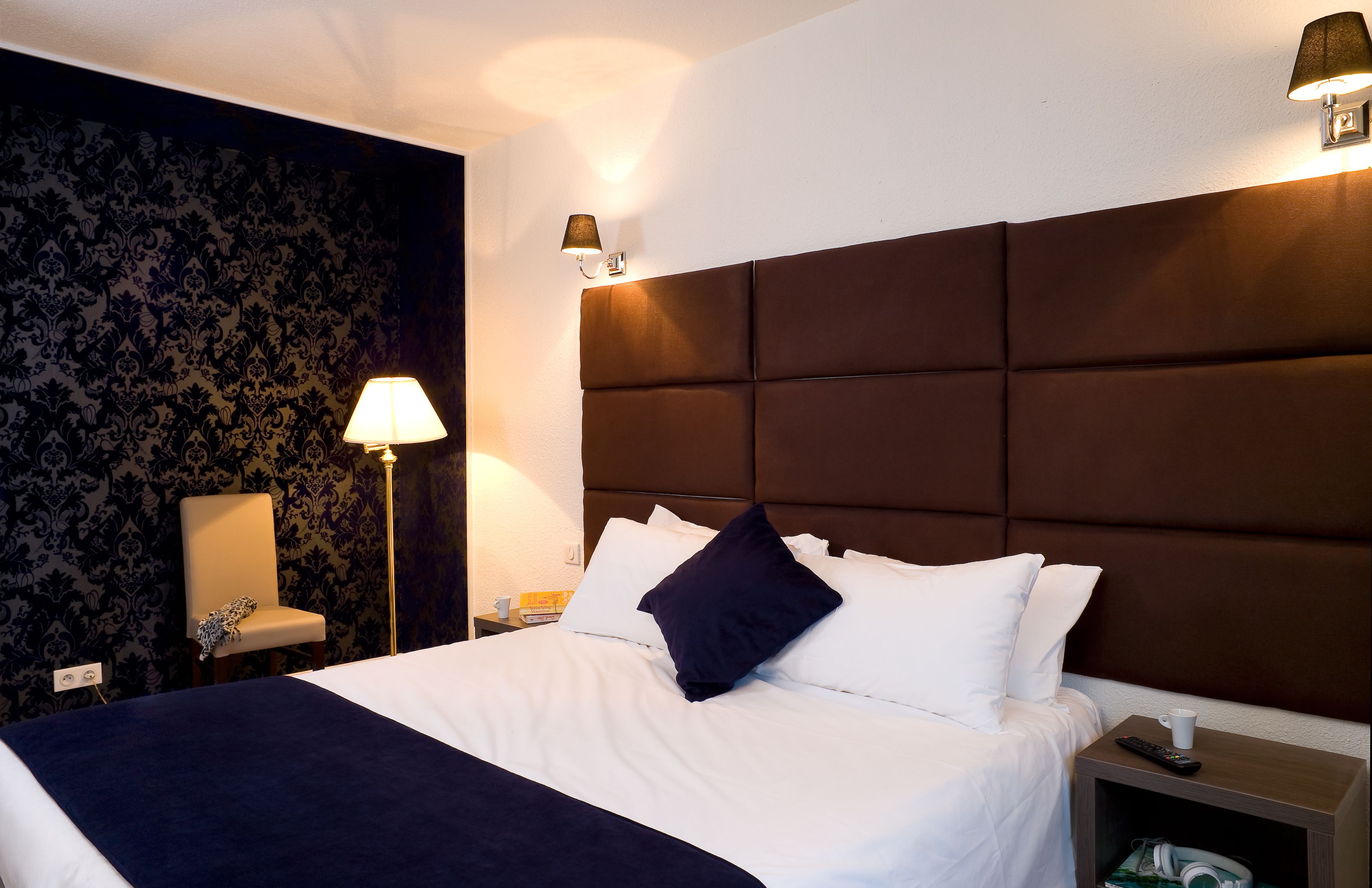 A neatly made bed with white linens and a dark blue throw blanket, a bedside lamp, a chair, and a nightstand with a remote control and a cup.