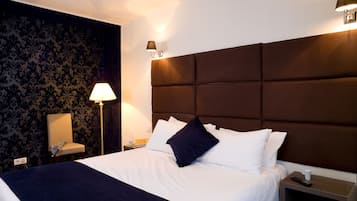 Una cama bien hecha con sábanas blancas y un edredón azul oscuro, una lámpara de noche, una silla y una mesita de noche con un control remoto y una taza.