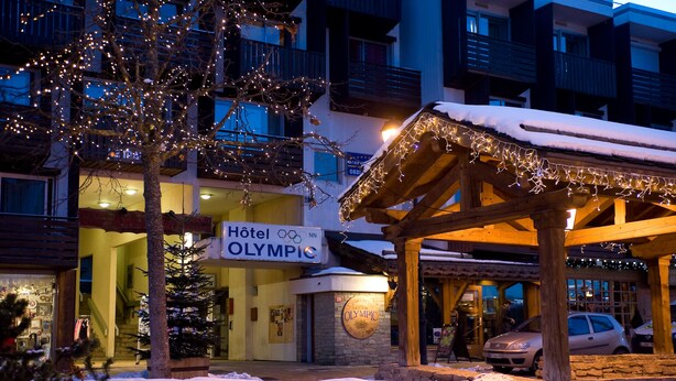 Un hotel con un cartel que dice 'Hotel Olympic', un área exterior cubierta con luces y nieve en el suelo.