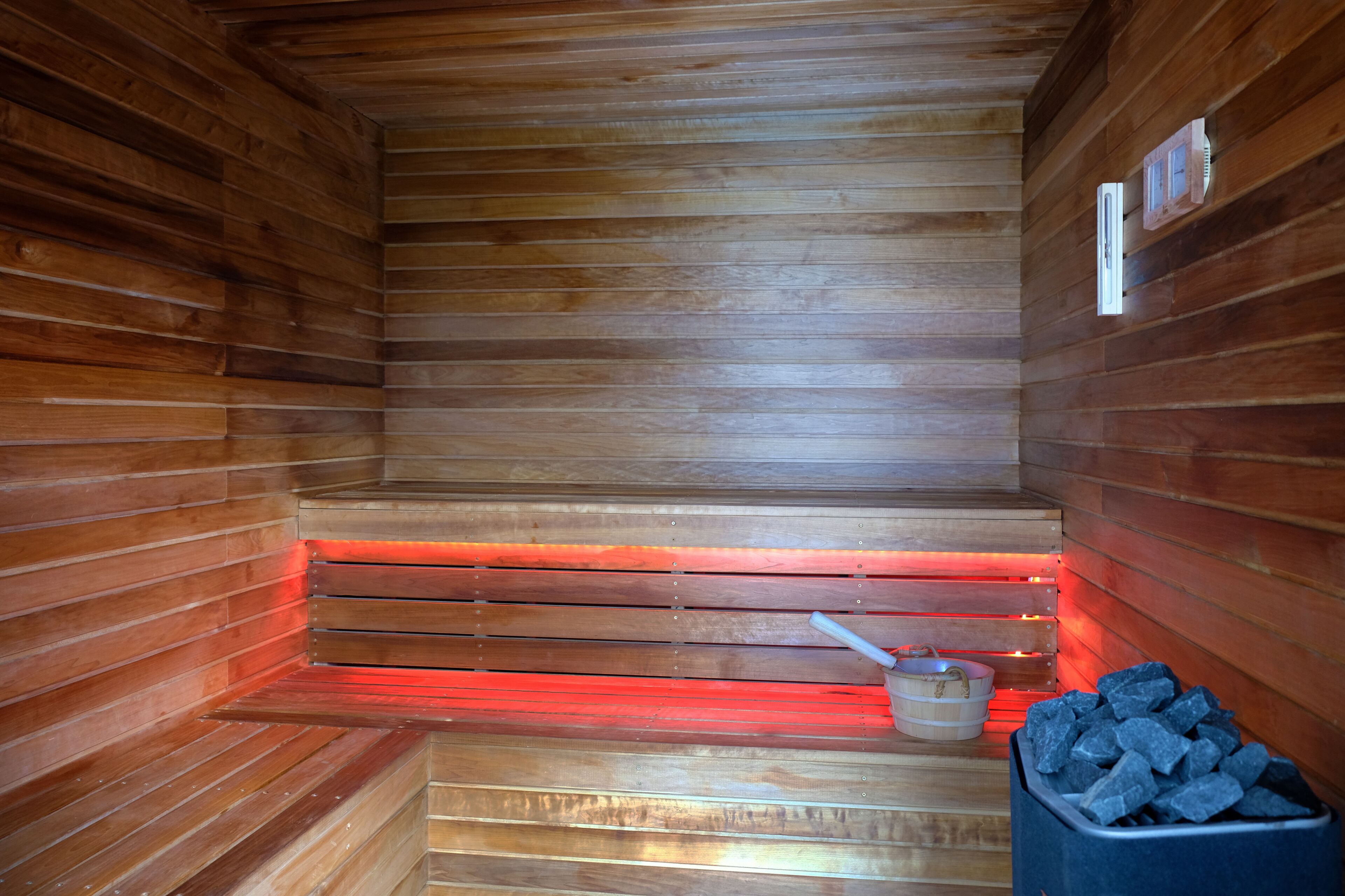 Un sauna aux murs et aux bancs en bois, agrémenté d’une bande lumineuse rouge et d’un seau rempli de pierres.