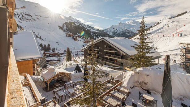 Un complejo turístico en la montaña cubierto de nieve, con edificios de madera, mesas al aire libre y pistas de esquí al fondo.