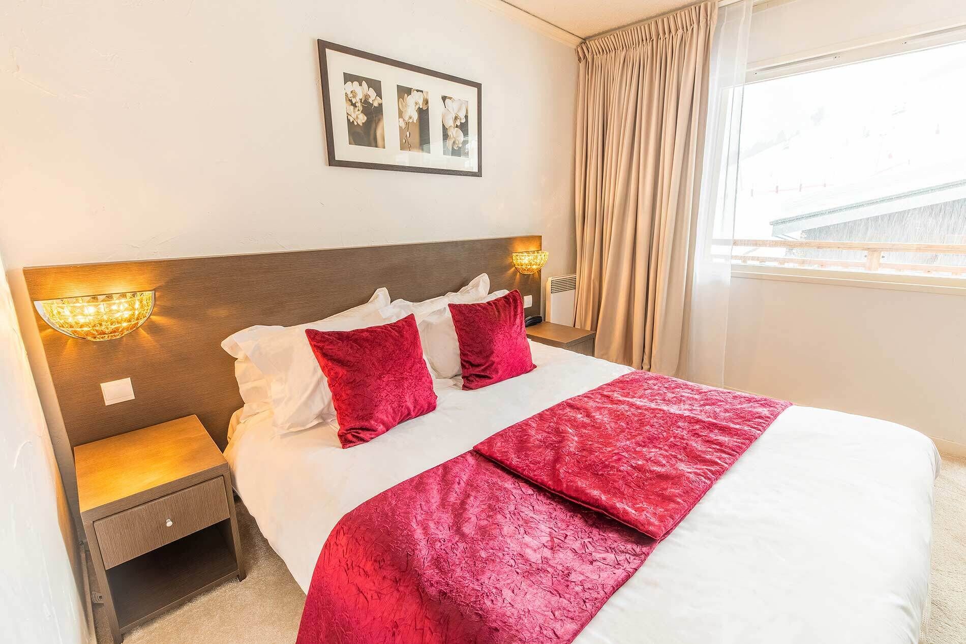 A hotel room with a large bed, two red pillows, a white bedspread, a wooden headboard, two bedside lamps, a nightstand, and a window with curtains.