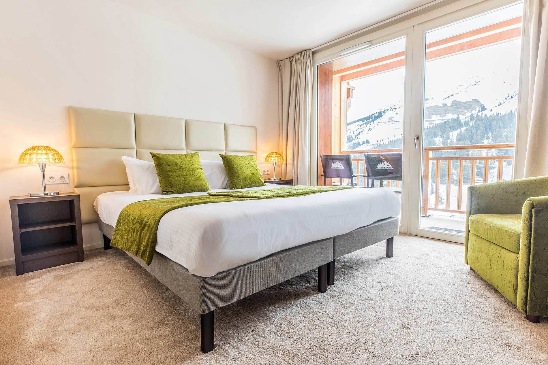 Une chambre d’hôtel avec un lit, deux tables de chevet, un fauteuil vert et un balcon offrant une vue sur la montagne.