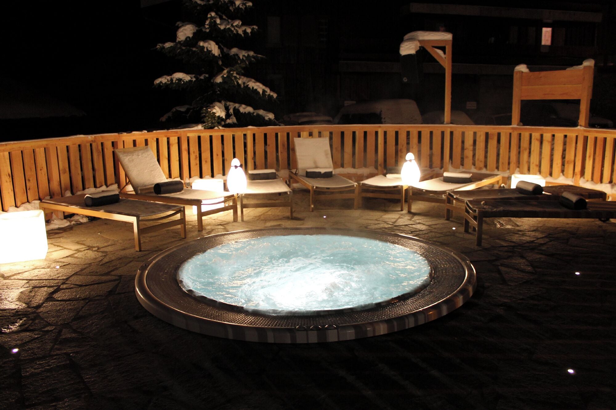 A hot tub surrounded by wooden lounge chairs with cushions, set on a stone patio at night.
