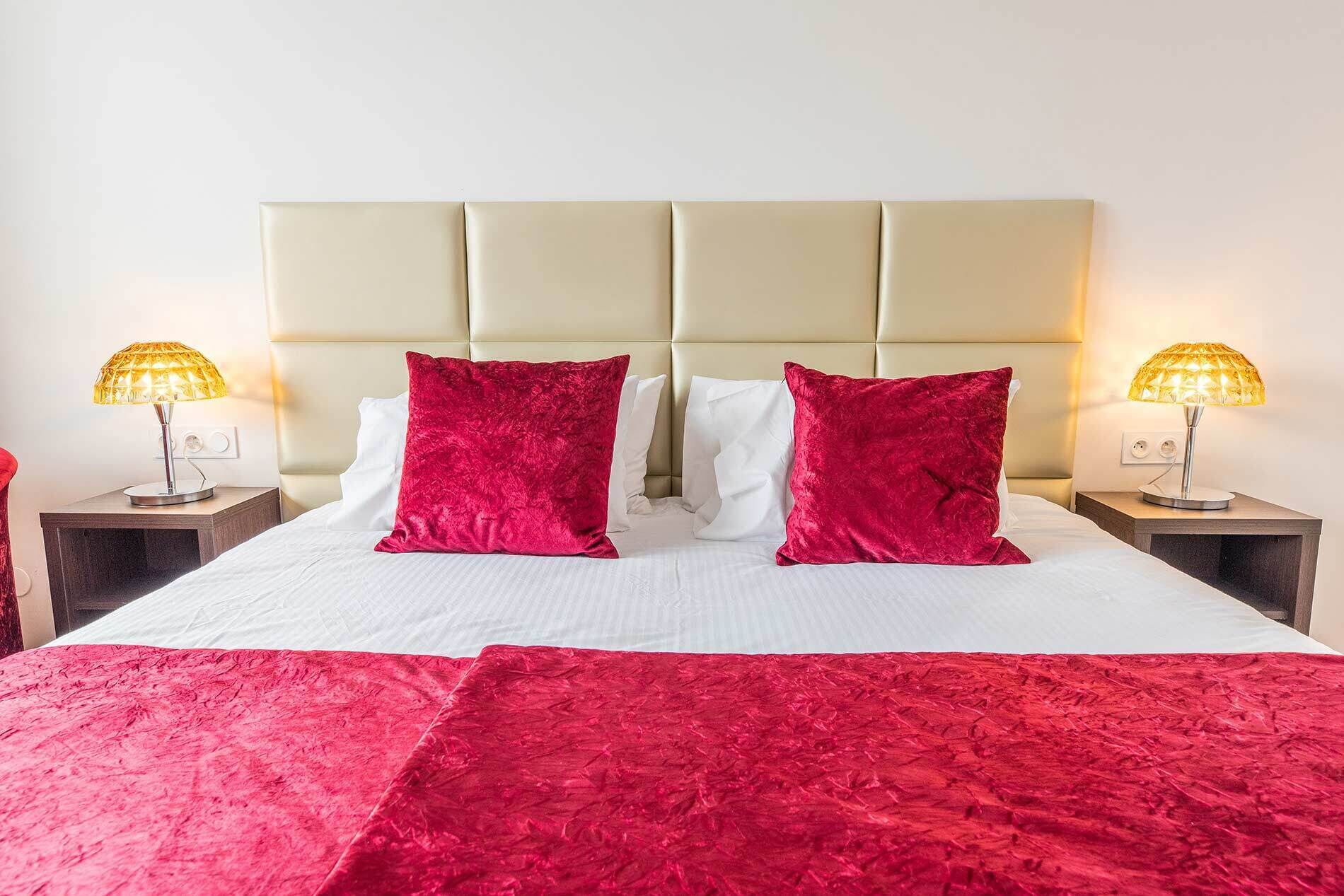 A bed with a beige headboard, two red pillows, and a red bedspread. There are two bedside lamps with yellow shades.