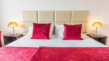 A bed with a beige headboard, two red pillows, and a red bedspread. There are two bedside lamps with yellow shades.
