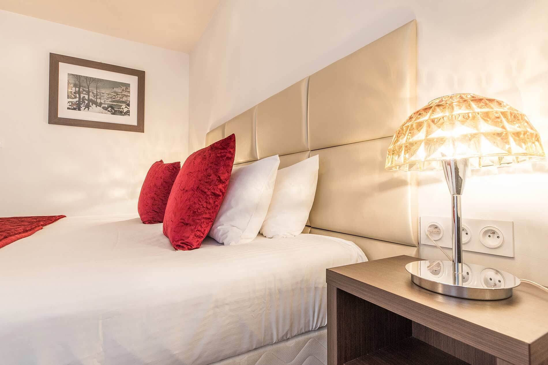Una cama bien hecha con almohadas blancas y rojas, una lámpara de noche y un cuadro en la pared.