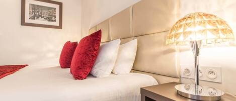 Una cama bien hecha con almohadas blancas y rojas, una lámpara de noche y un cuadro en la pared.