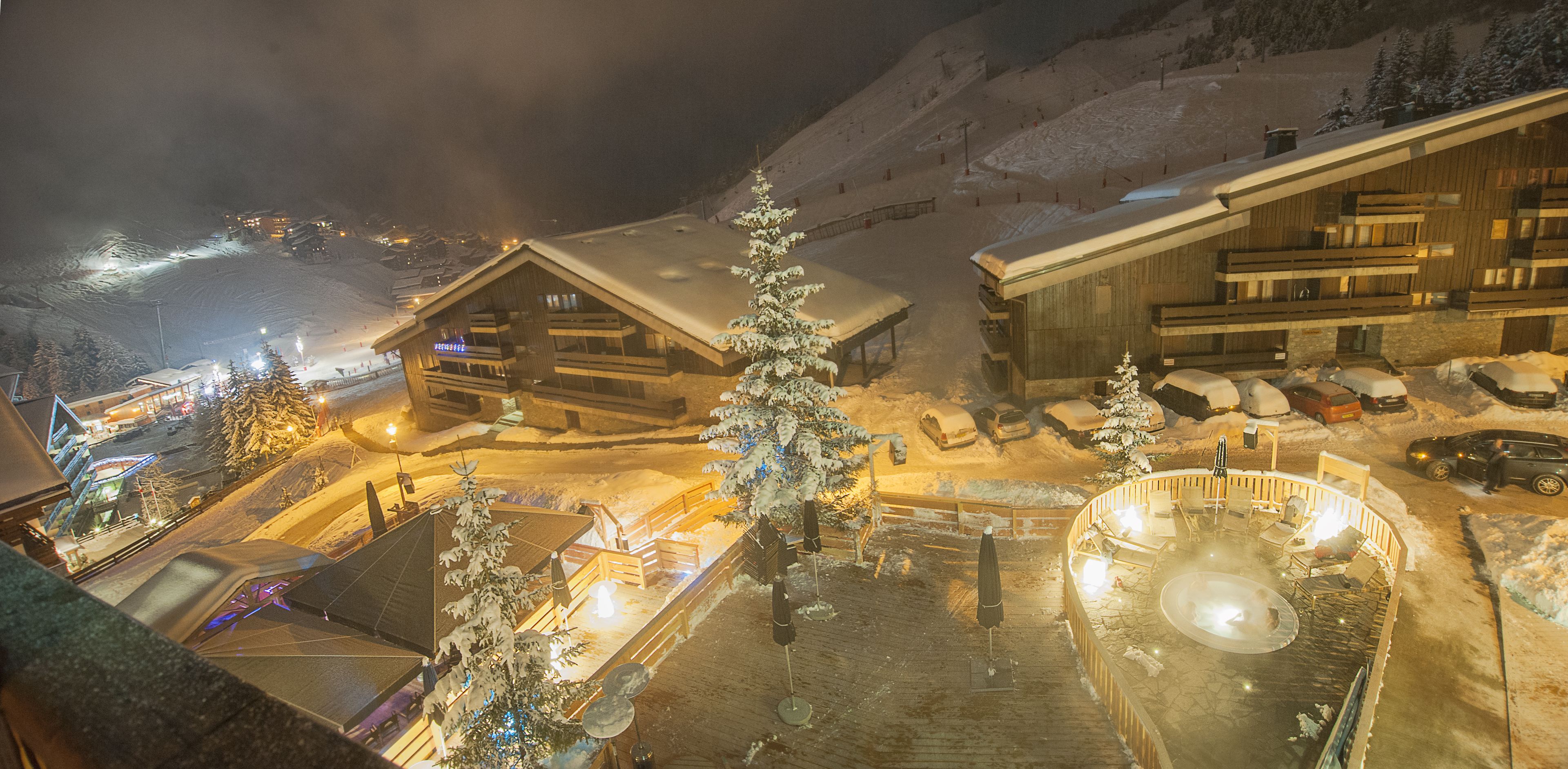 A snow-covered lodge with a ski lift in the background, a heated outdoor seating area, and a wooden building with a sloped roof.