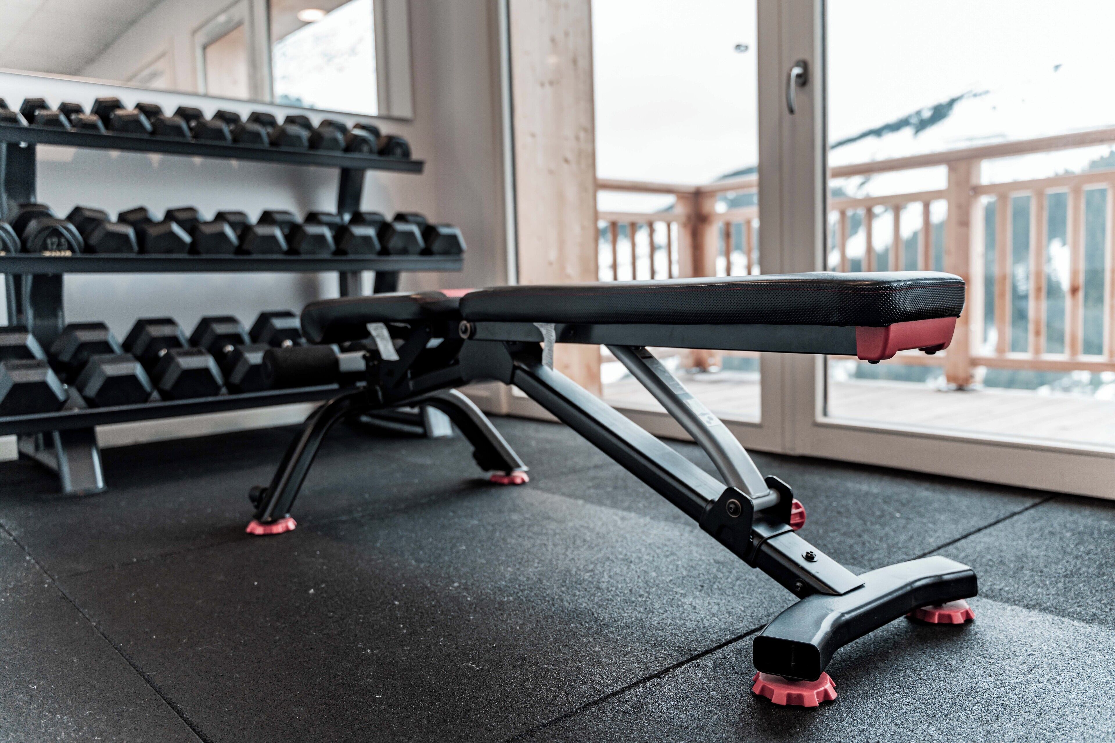 A gym with a bench press, dumbbells, and a view of a wooden deck outside.