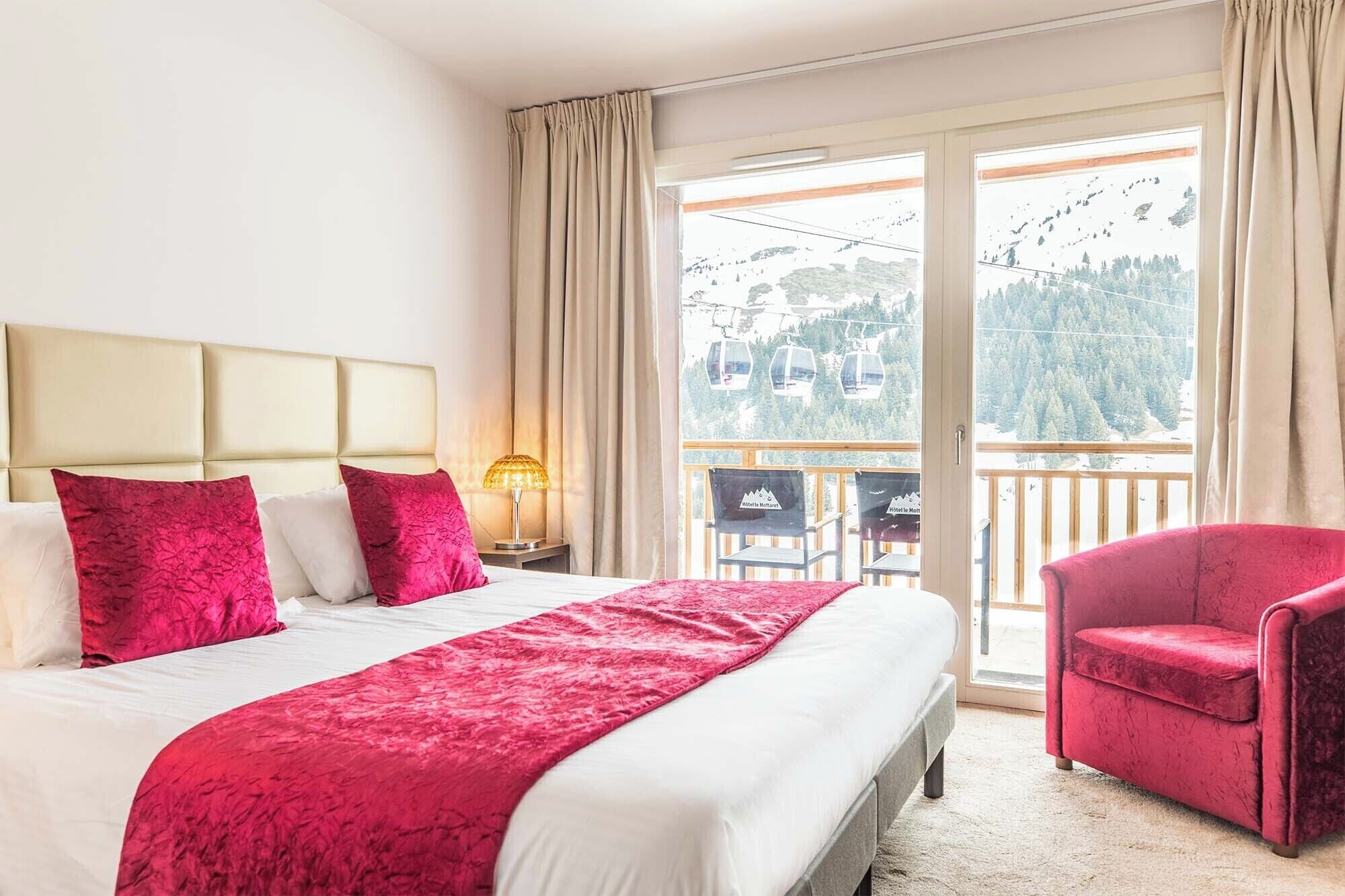 A hotel room with a large bed, a red chair, and a balcony with a mountain view.