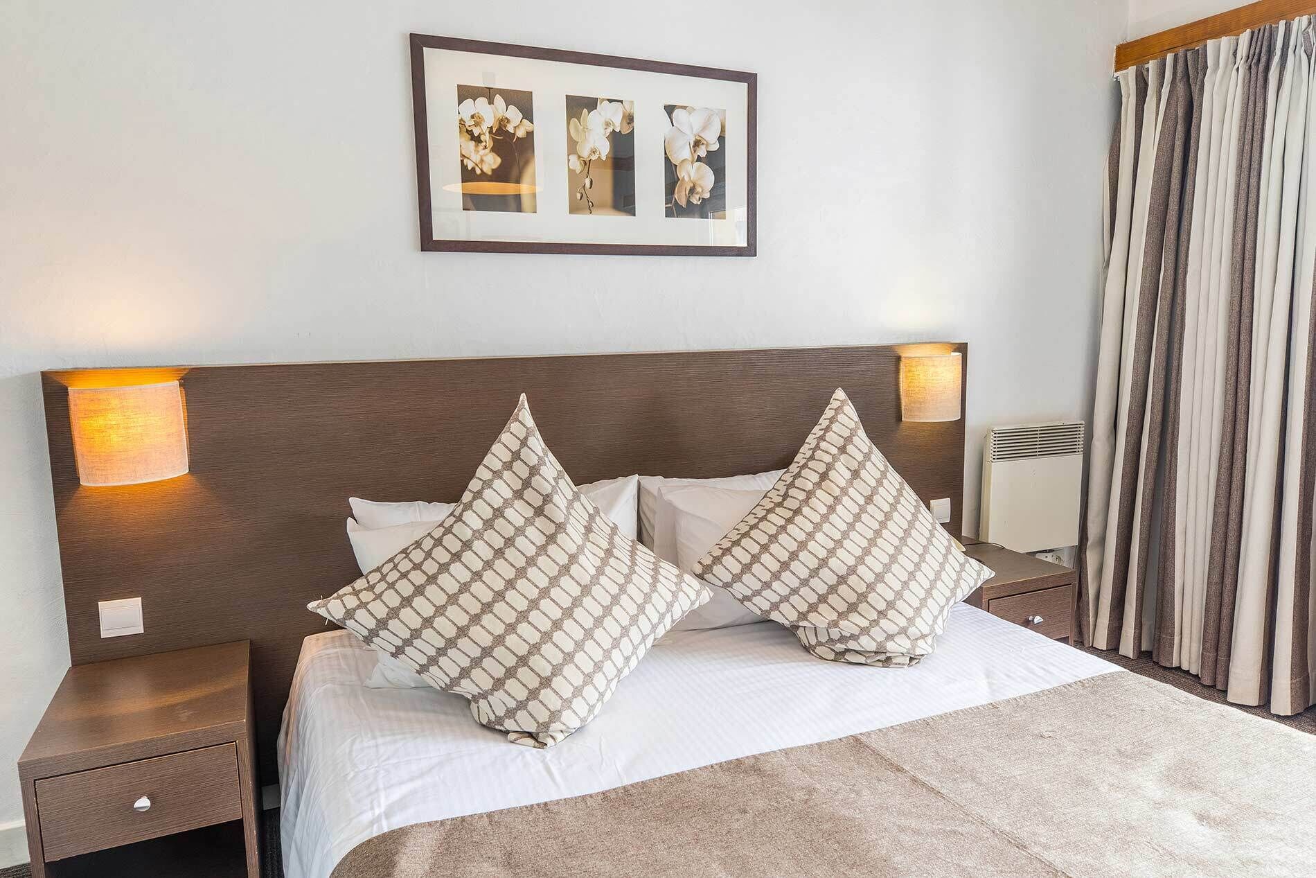 Una cama bien hecha con dos almohadones estampados, un cabecero con lámparas fijadas a la pared y un cuadro enmarcado encima.