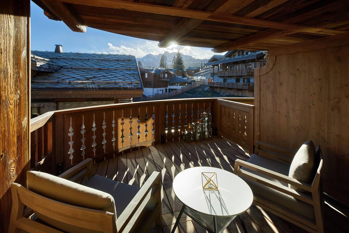 Een houten balkon met een ronde tafel en twee stoelen, met uitzicht op een bergachtig dorp met traditionele gebouwen.