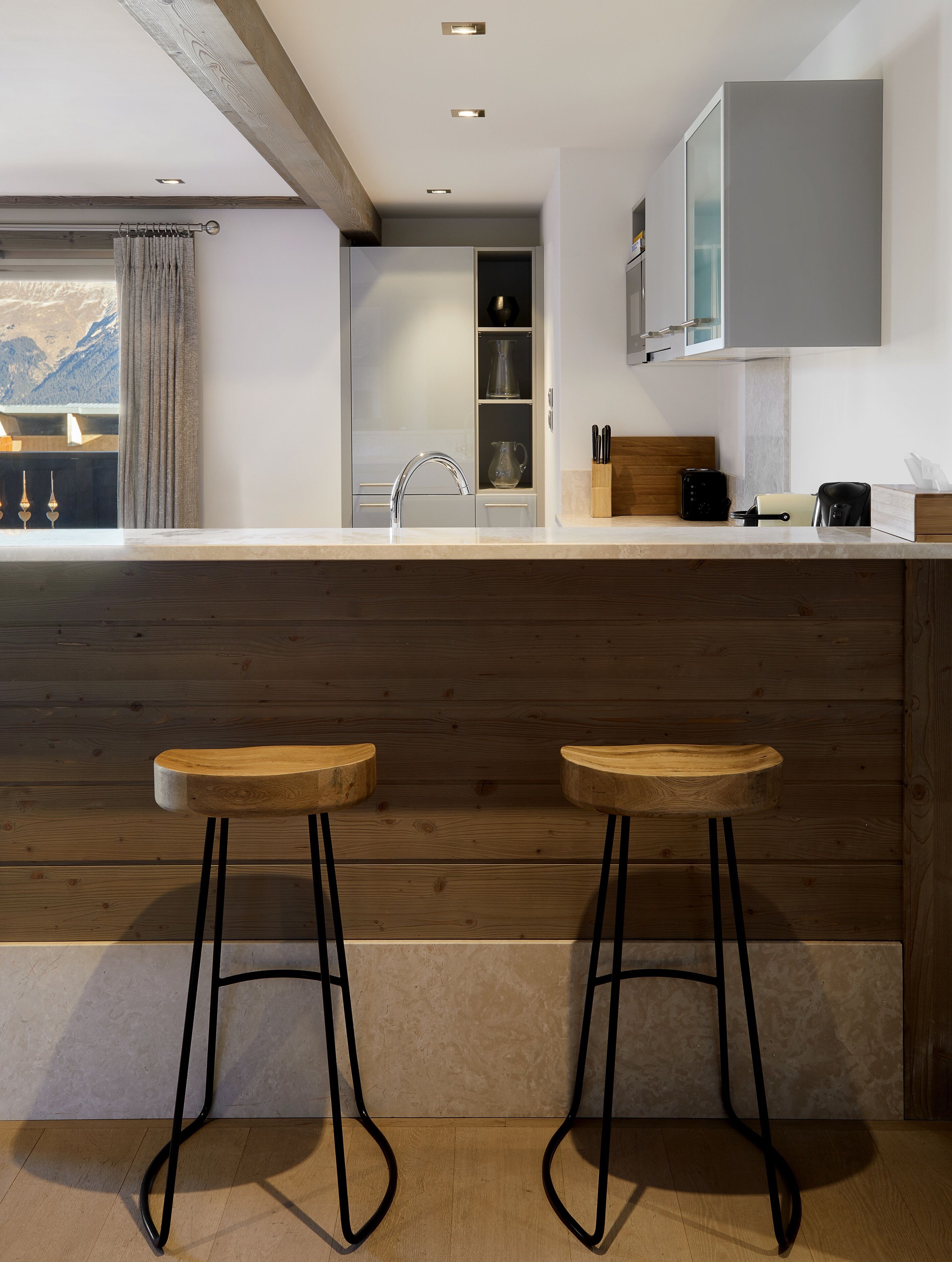 A modern kitchen with a wooden bar area, two stools, and a view of mountains through a window.