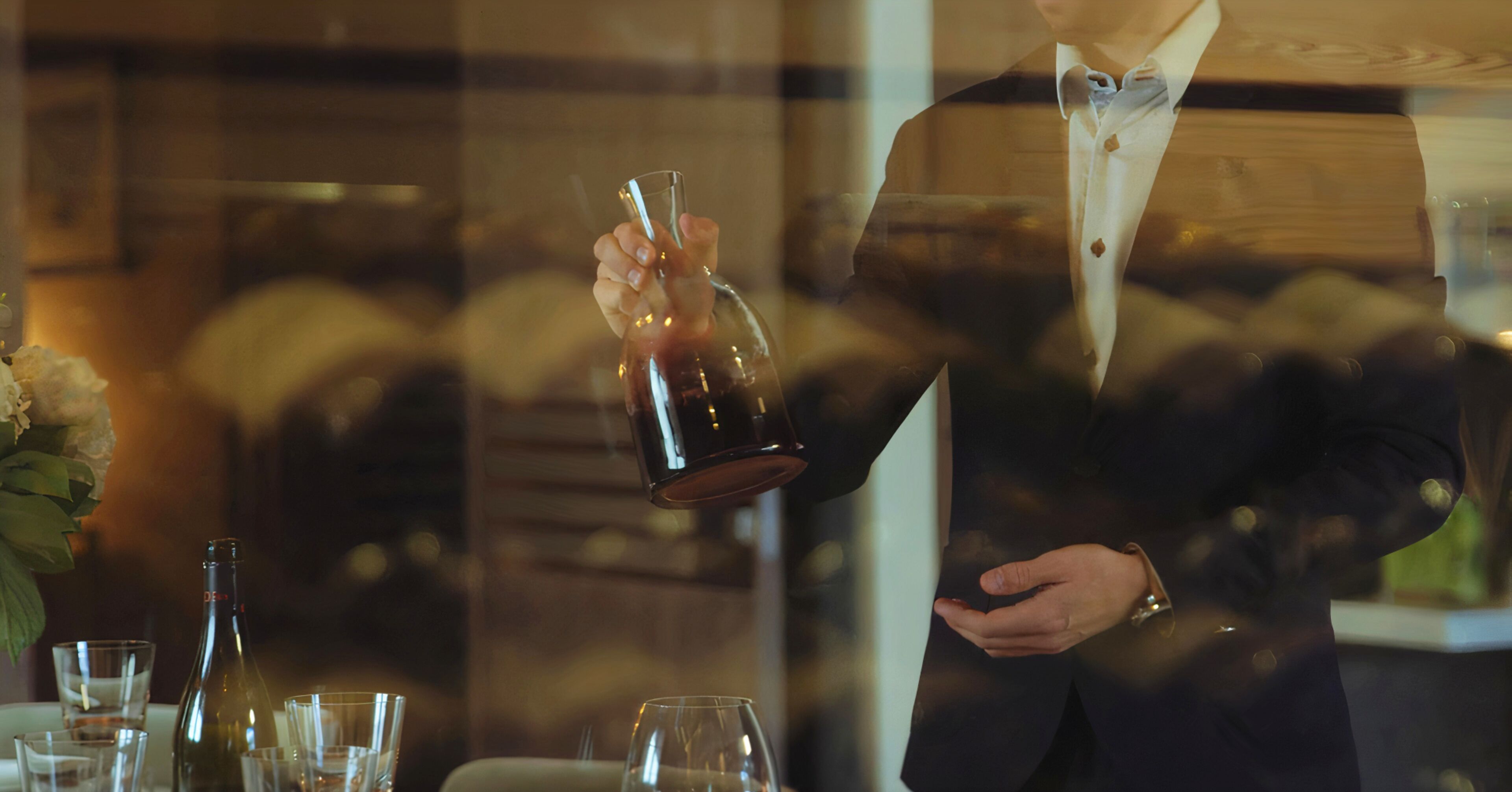 Une personne verse une boisson dans un verre, sur une table où se trouvent d’autres verres et une bouteille.