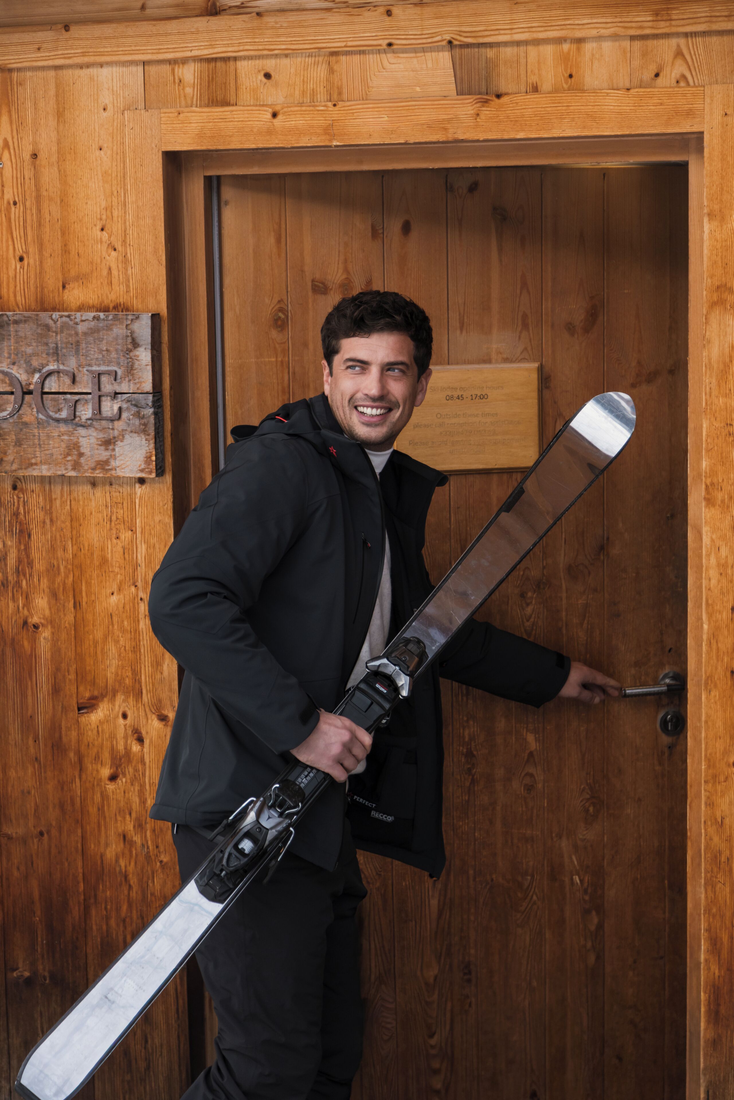 A person holding a ski board, standing in front of a wooden door with a sign that reads 'OGI'.