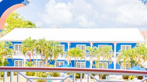 A balcony with white chairs and a table, overlooking a pool and a blue building with palm trees. - Bay Gardens Inn (Gros Islet)