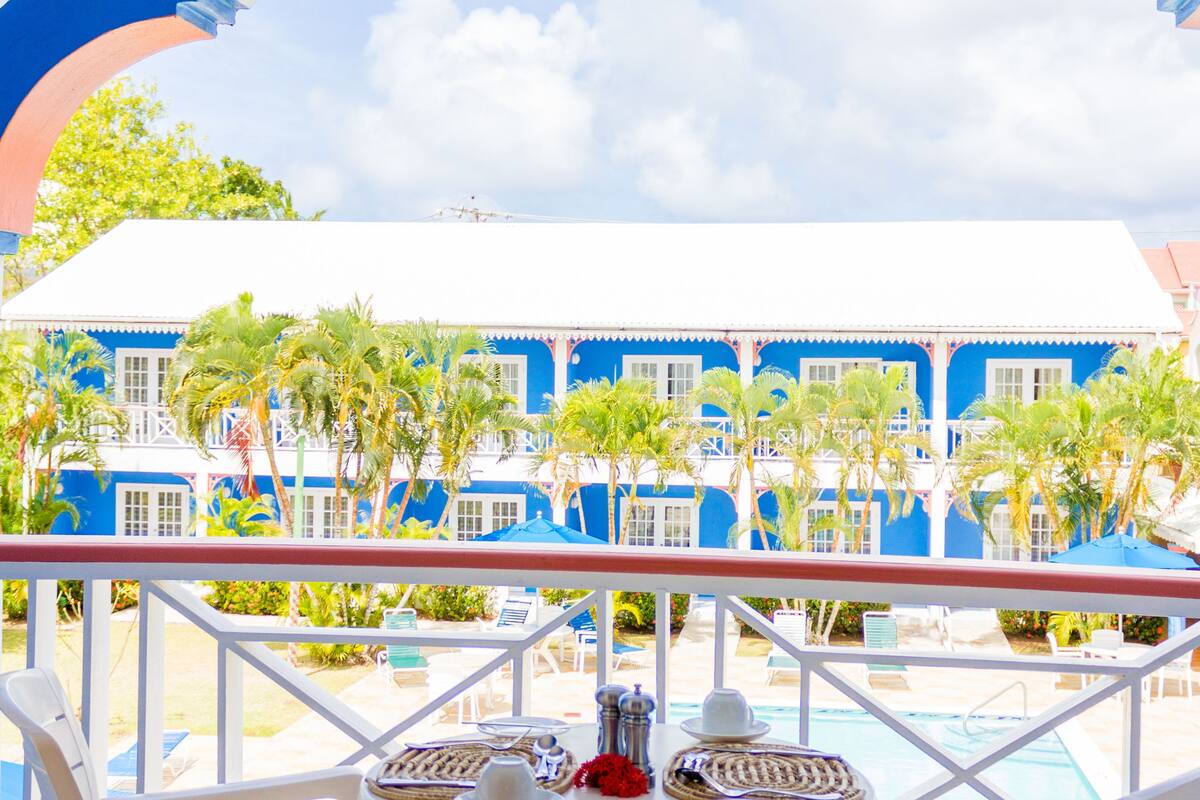A balcony with white chairs and a table, overlooking a pool and a blue building with palm trees.