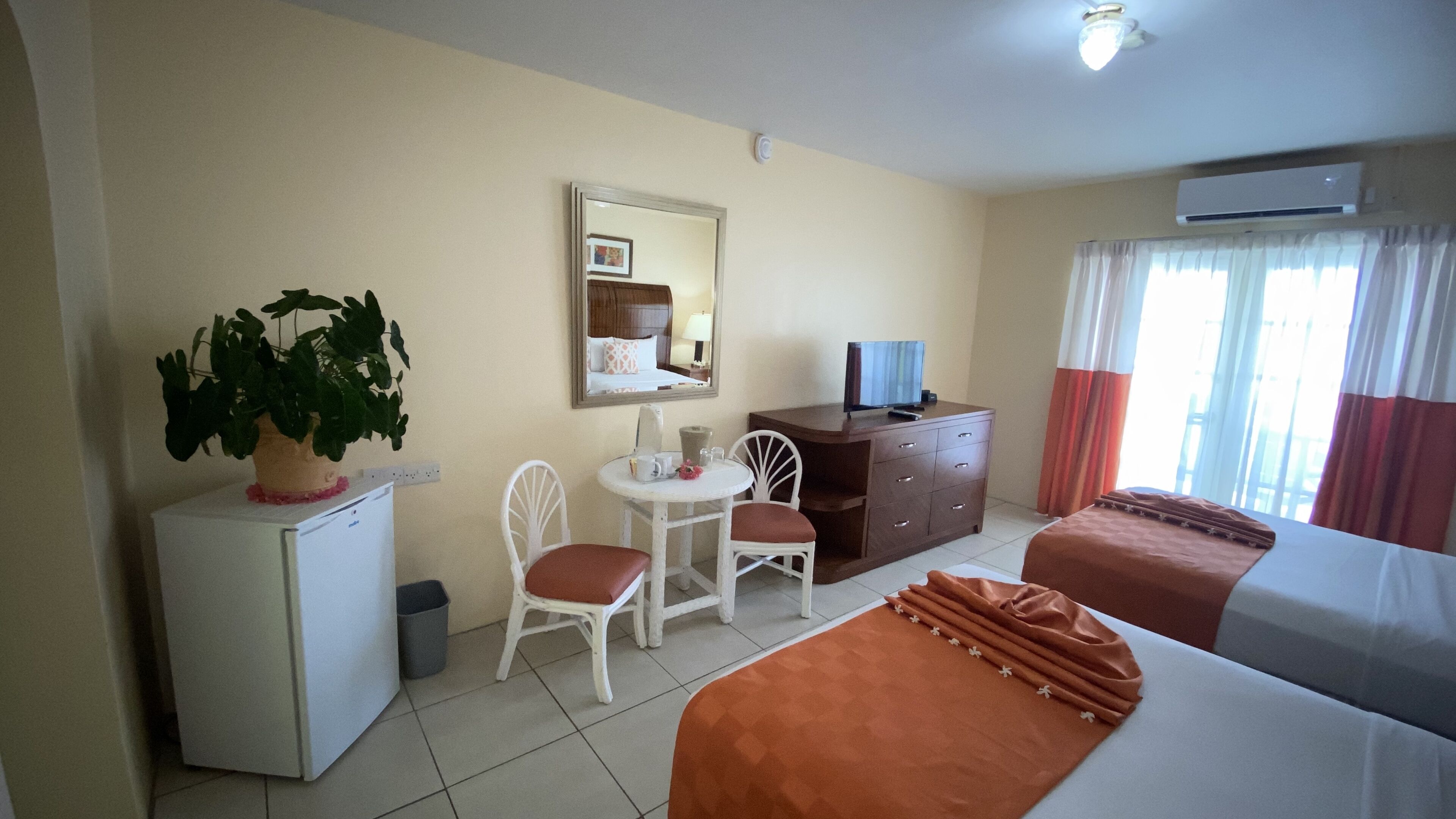 A hotel room with a bed, a desk with a chair, a small refrigerator, a potted plant, a mirror, and a window with curtains.