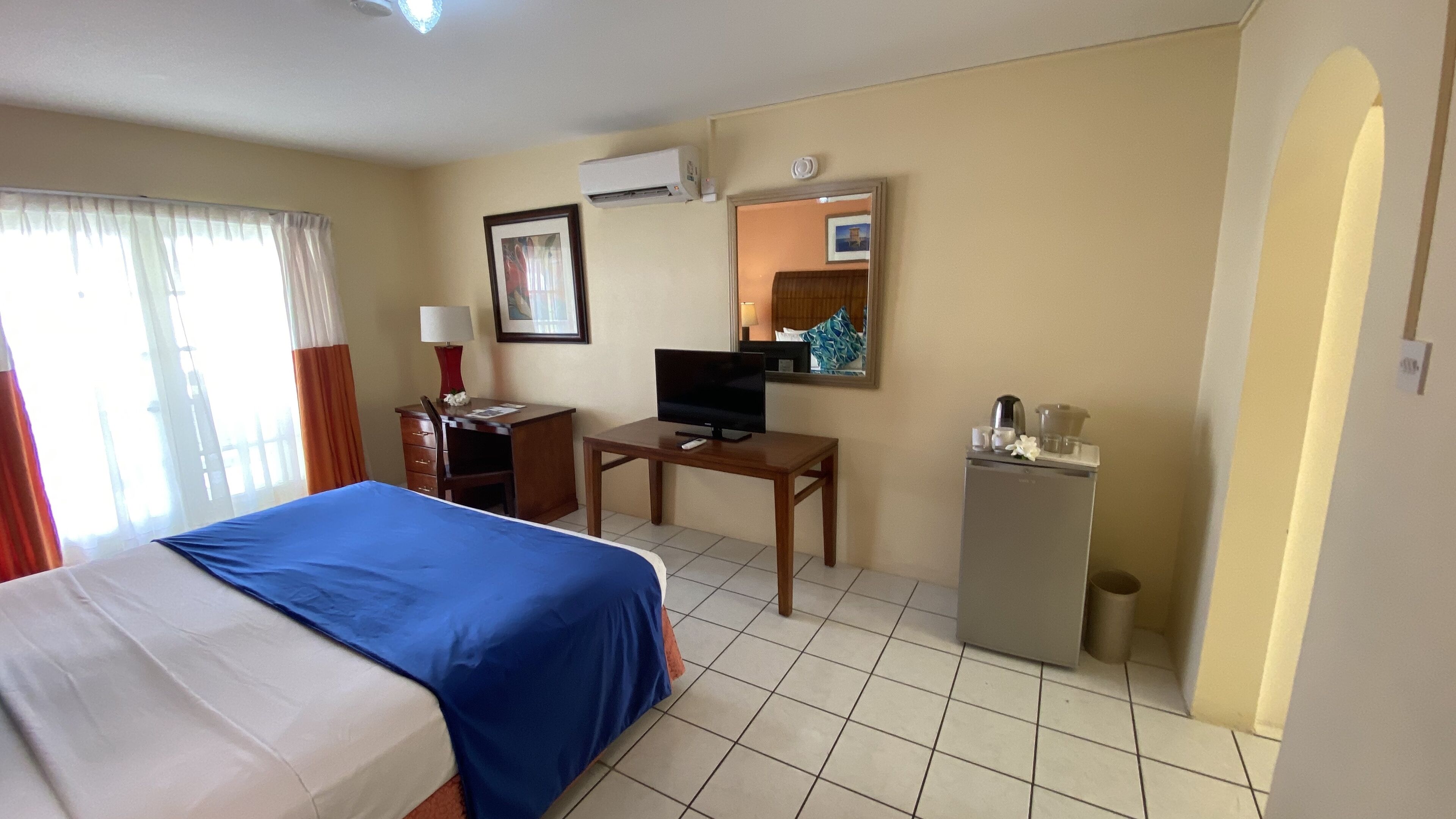Habitación de hotel con cama, escritorio con televisor, refrigerador pequeño y ventana con cortinas.