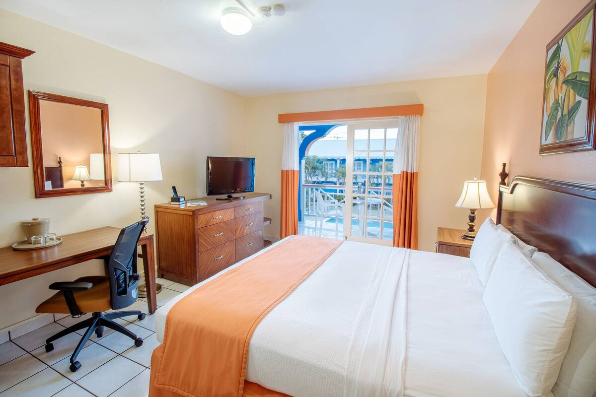 A hotel room with a bed, desk, chair, television, and a view of a pool and building through the window.
