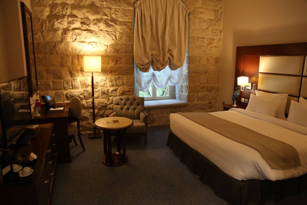 Une chambre d’hôtel avec un mur en pierre, un grand lit, un bureau avec une chaise, une table ronde et une fenêtre avec des rideaux.