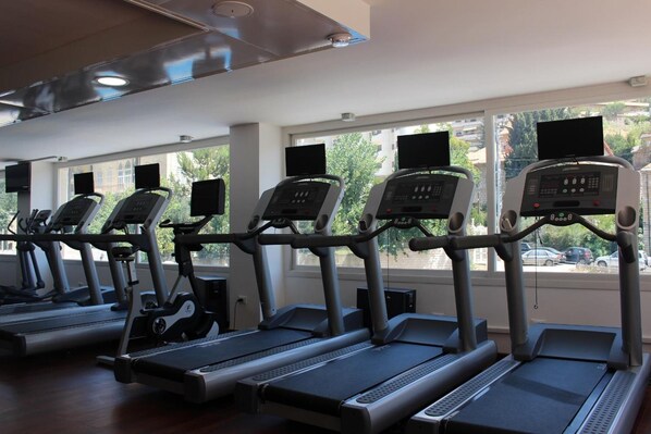 A row of stationary bikes with digital displays, positioned in a gym with large windows and a view of the outside. - Grand Kadri Hotel (Zahlé)