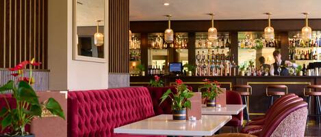 A bar area with red upholstered seating, a bar counter stocked with bottles, and hanging pendant lights.