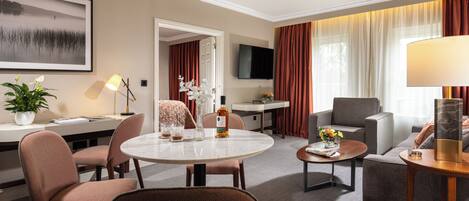 A hotel room with a dining area, a sofa, a TV, and a large window with curtains.