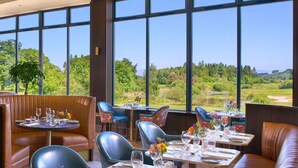 Un restaurante con grandes ventanales que ofrecen vistas a un paisaje verde. Las mesas están dispuestas con copas de vino y platos.