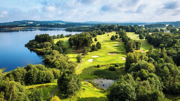 Un campo de golf con un lago, rodeado de árboles frondosos y ondulantes colinas.