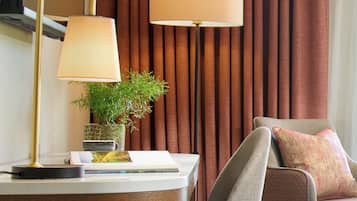 A hotel room with a desk, a lamp, a chair, a potted plant, and a large window with curtains.