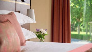 A neatly made bed with white and pink pillows, a vase with flowers, and a view of greenery outside.