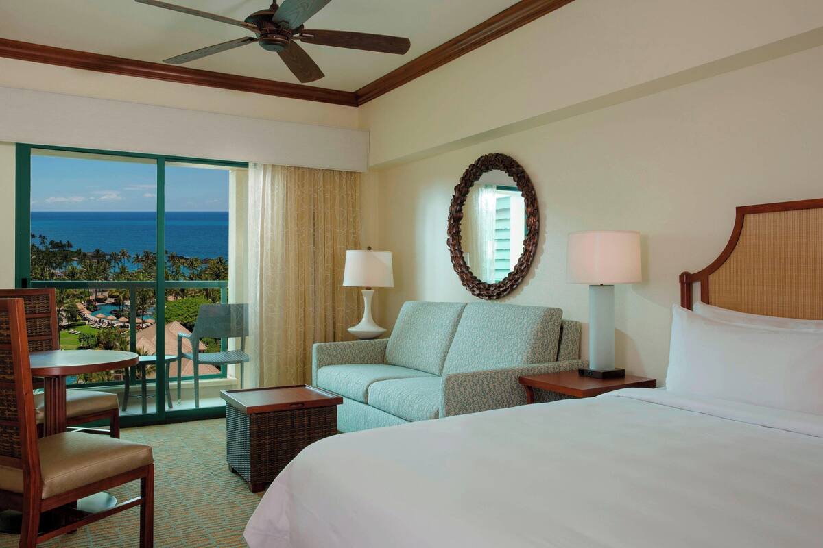 A hotel room with a bed, a sofa, a chair, a small table, a lamp, a mirror, and a view of the ocean through large windows.