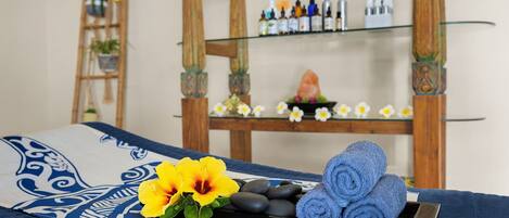 Una cama con colcha azul y blanca, una bandeja con una flor amarilla, toallas azules y un estante de madera con botellas y un objeto decorativo al fondo.