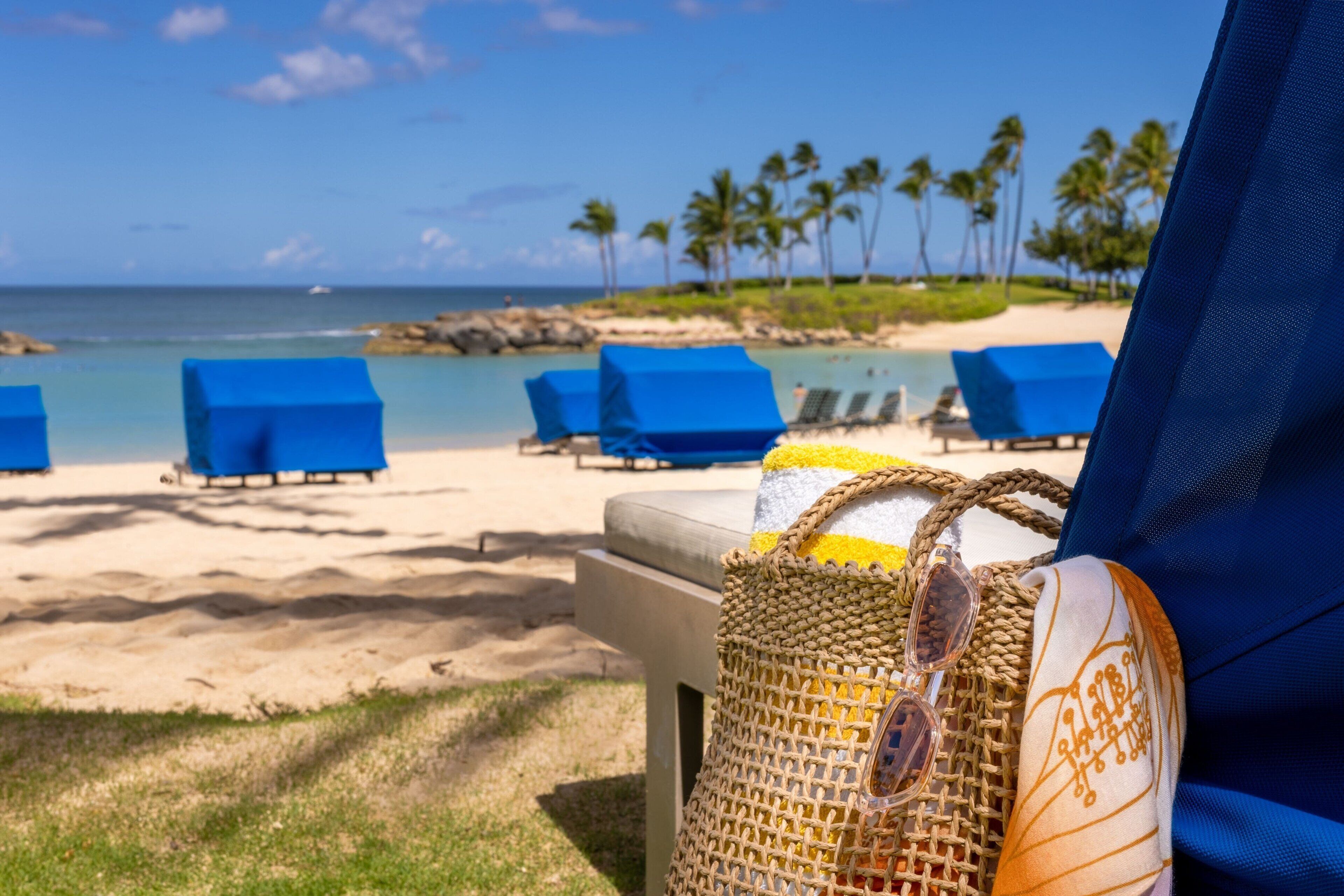 A beach view with blue lounge chairs, a straw bag with sunglasses, and a clear sky.
