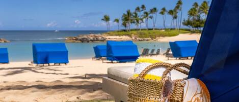 A beach view with blue lounge chairs, a straw bag with sunglasses, and a clear sky.