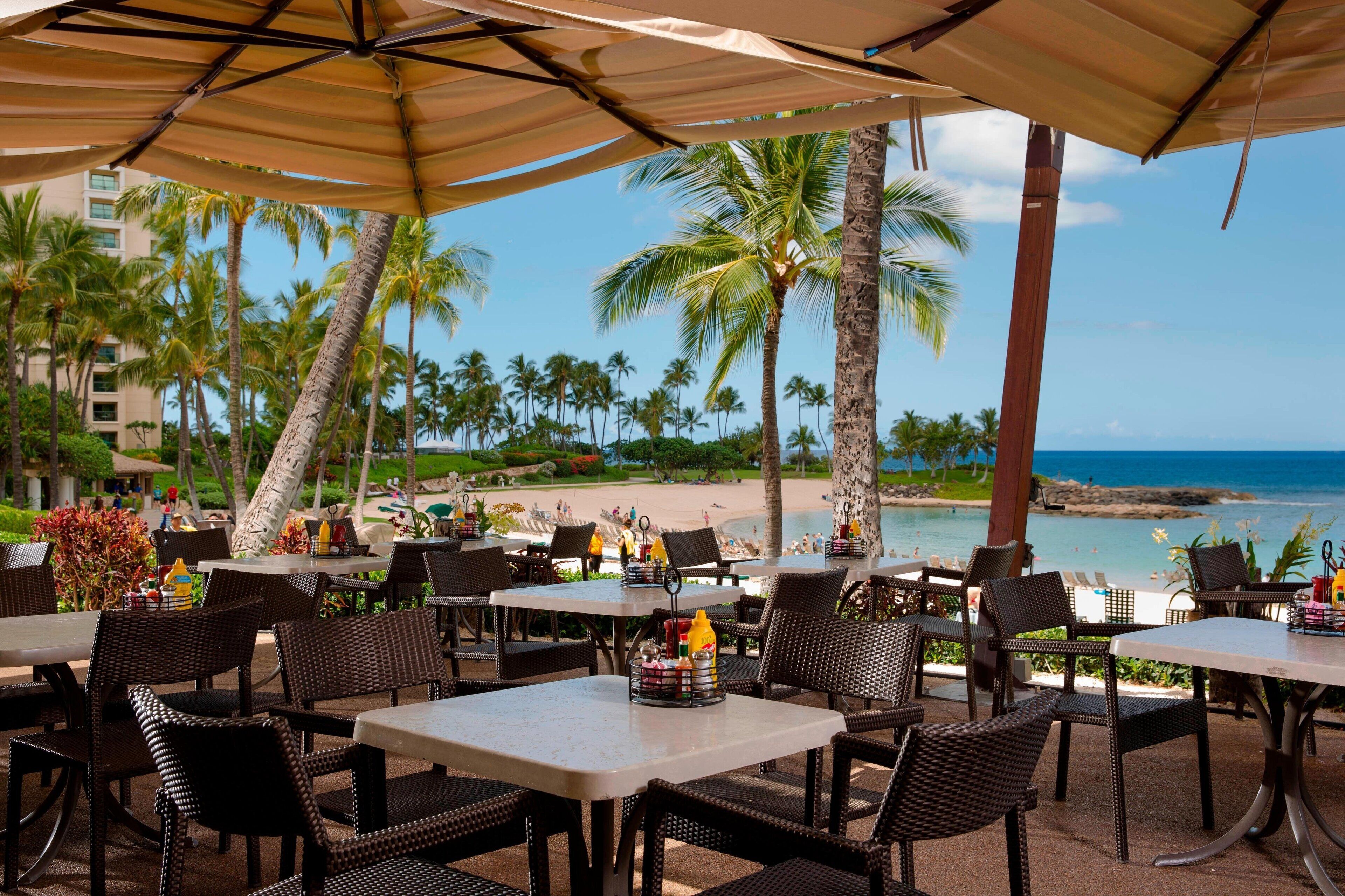 Un área de comedor al aire libre con mesas y sillas, con vista a una playa y al océano.