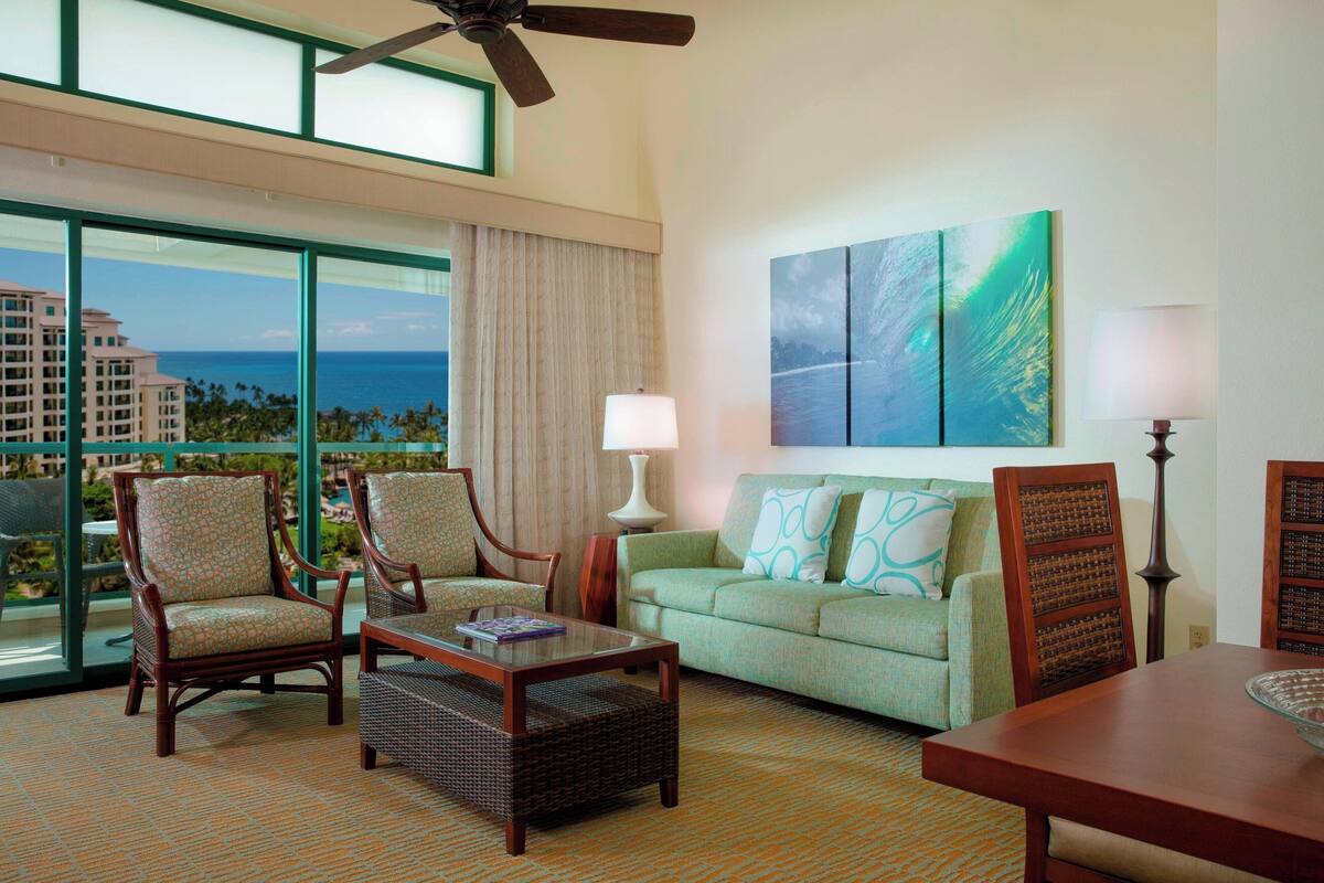 A hotel room with a large window offering a view of the ocean and buildings, a green sofa with patterned cushions, two wooden chairs, a small table, and a ceiling fan.
