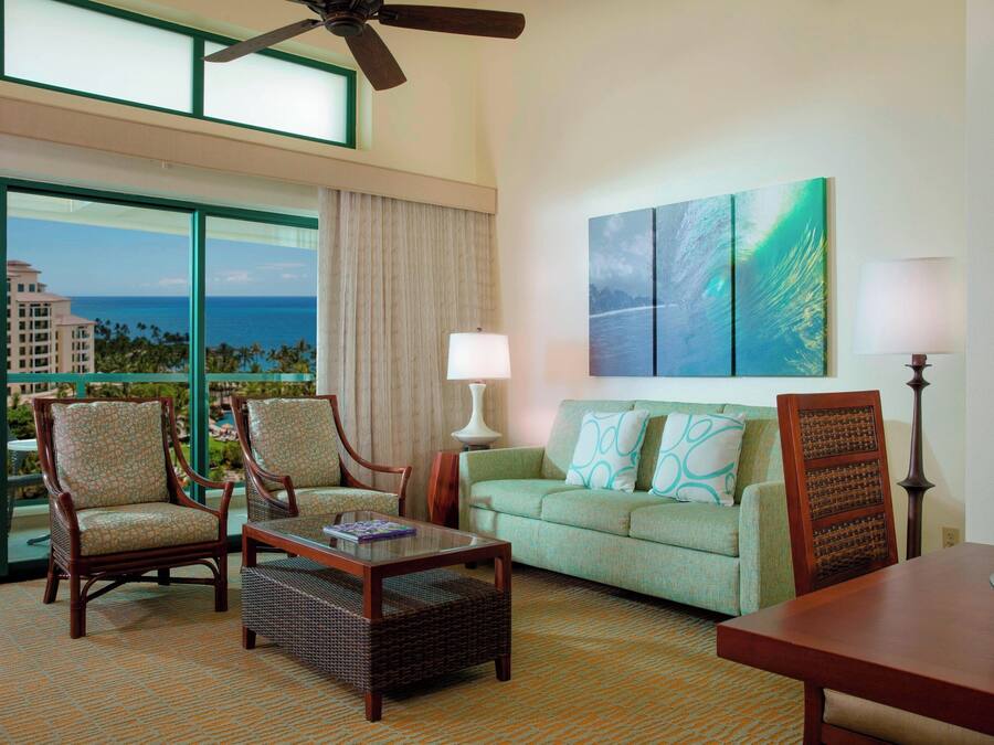 A hotel room with a large window offering a view of the ocean and buildings, a green sofa with patterned cushions, two wooden chairs, a small table, and a ceiling fan.