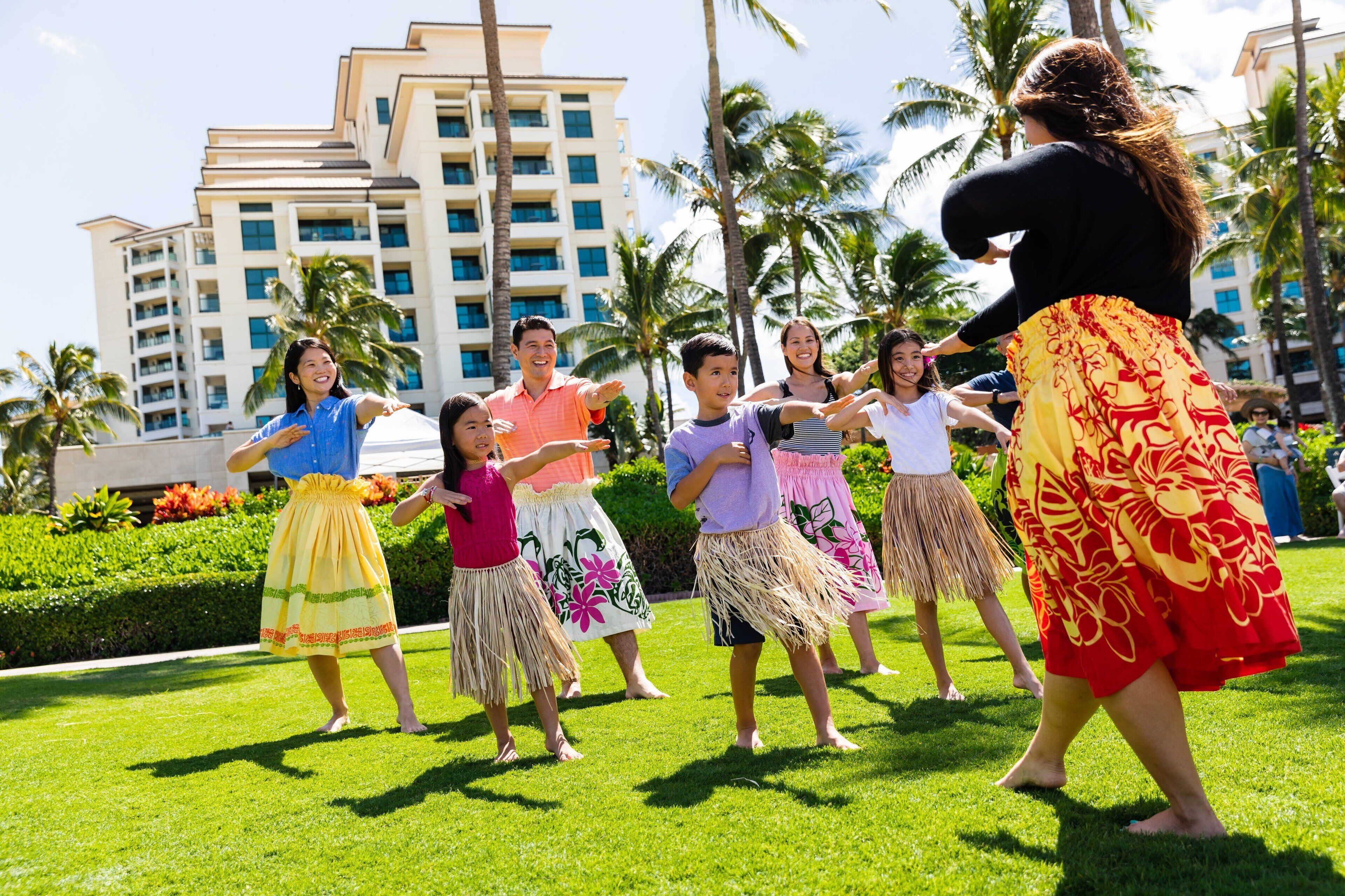 Un groupe de personnes, vêtues de costumes traditionnels, exécutent une danse sur une pelouse, avec un bâtiment d’hôtel en arrière-plan.