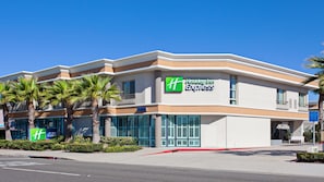 Edificio de Holiday Inn Express con un diseño moderno, que presenta un llamativo logotipo en forma de 'H', amplias ventanas y palmeras al frente.