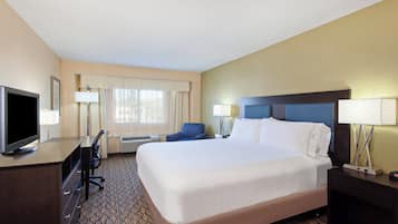 A hotel room with a bed, a desk with a TV, a chair, a desk lamp, and a bedside table with a lamp.