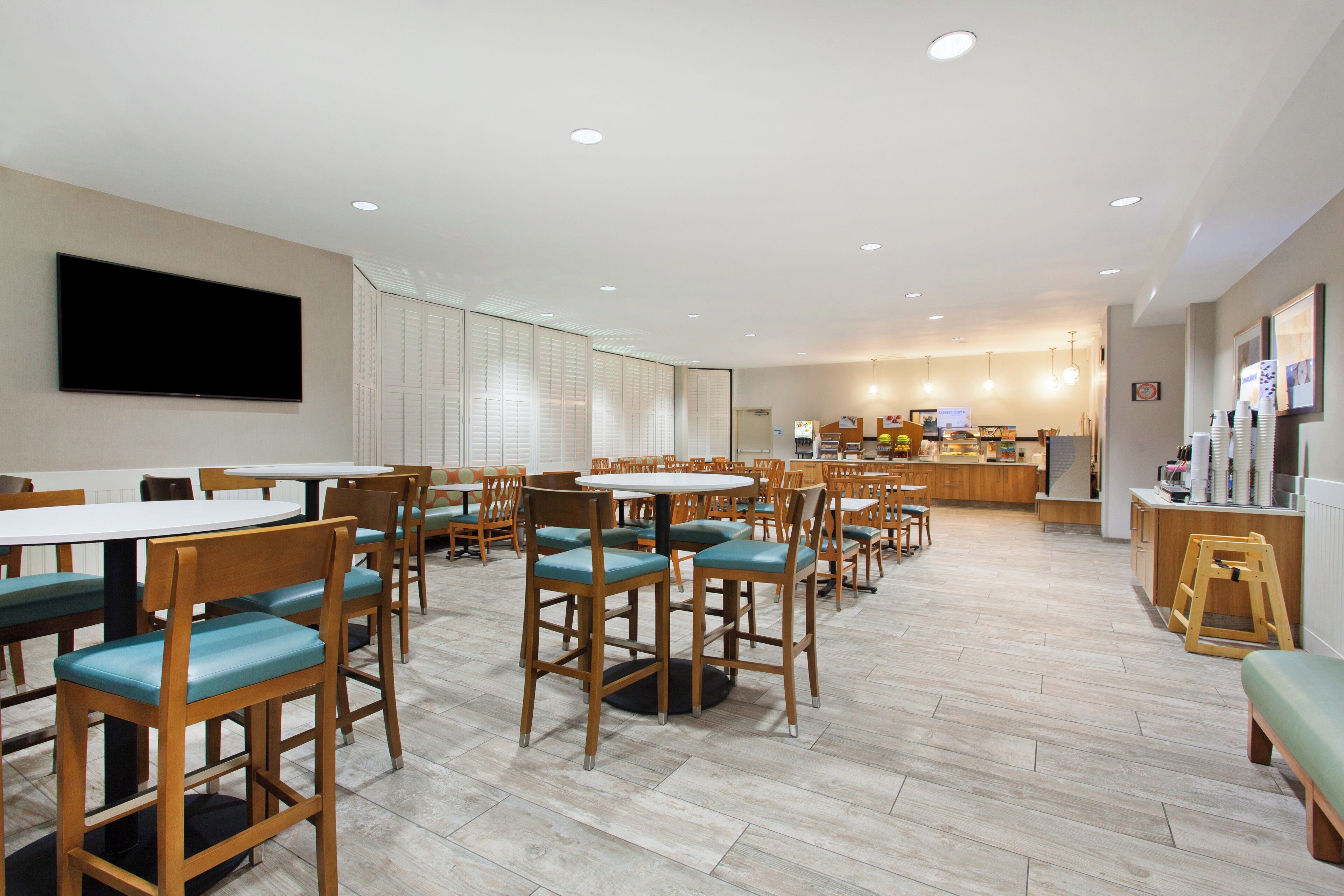 A hotel dining area with multiple tables and chairs, a flat-screen TV mounted on the wall, and a kitchen area in the background.