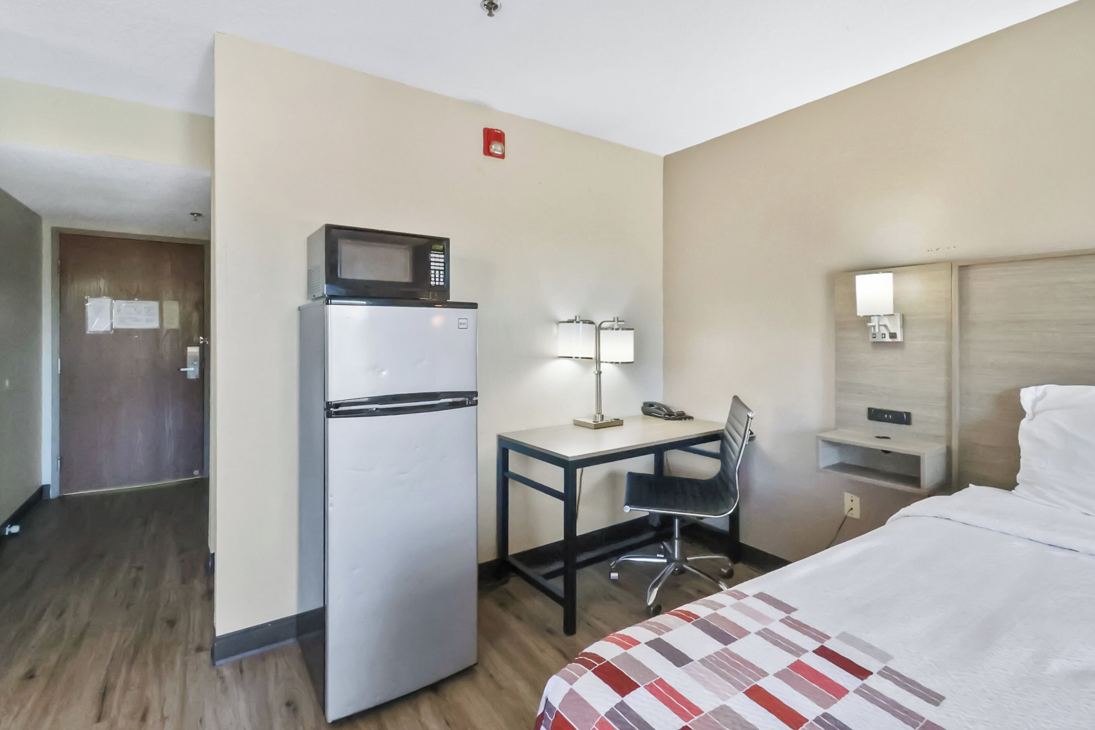 Habitación de hotel con cama, escritorio con silla, refrigerador, microondas, lámpara, teléfono y mesita de noche.